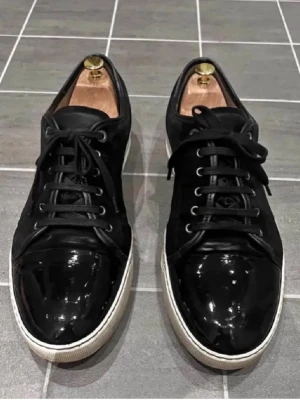 Svarta Lanvin sneakers i lack och mocka - Snygga svarta sneakers från Lanvin med en mix av glansigt lack och mjuk mocka. Skorna har vita sulor och svarta skosnören för en clean look. Perfekta för dig som gillar exklusiva och stilrena sneakers med en lyxig touch. pris kan diskuteras vid snabb affär