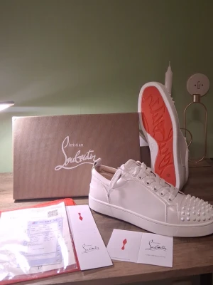 Vita Christian Louboutin sneakers - Säljer ett par vita sneakers från Christian Louboutin med klassisk röd sula och coola nitar på tån. Skorna är låga och har vita skosnören samt detaljer i vitt läder. Perfekta för dig som vill sticka ut med exklusiv design och edgy vibe. Kommer med allt du behöver😀