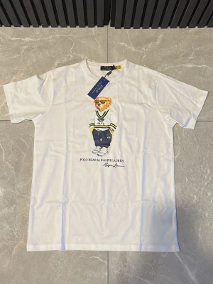 Vit Polo Bear t-shirt från Ralph Lauren - Vit t-shirt från Ralph Lauren med Polo Bear-tryck framtill. Klassisk passform med rund hals och korta ärmar. Tillverkad i mjuk bomull och har en stilren design med ikonisk björn i preppy-outfit.