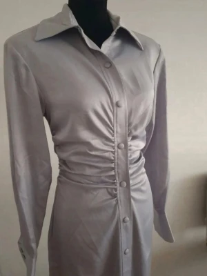 Långklänning - Stilren ljusgrå klänning med långa ärmar och markerad midja med rynkdetaljer. Klänningen har klassisk krage och knäppning framtill hela vägen ner. Materialet ser ut att vara glansigt, vilket ger en elegant känsla.