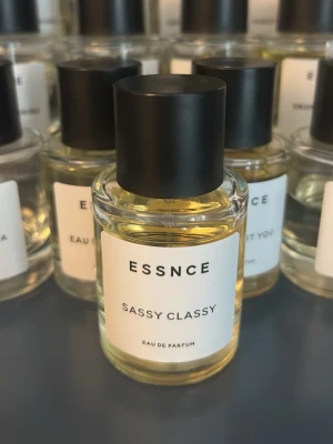 ESSNCE Sassy Classy Eau de Parfum - ESSNCE Sassy Classy Eau de Parfum i en stilren, rund glasflaska med svart matt kork. Parfymen har en ljusgul vätska och minimalistisk vit etikett med svart text. Perfekt för dig som gillar moderna och eleganta dofter.