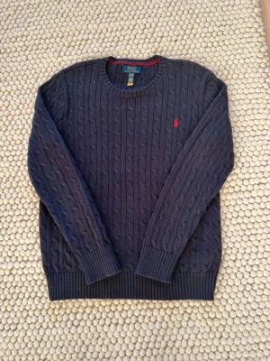  Polo Ralph Lauren blå tröja - Kabelstickad mörkblå tröja från Polo Ralph Lauren med rund halsringning och röd broderad logga på bröstet. Tröjan har ribbade muddar vid ärmslut och nederkant. Perfekt för dig som gillar klassisk och stilren design. OBS- det är XL i barnstorlek men passar M-L i vuxenstorlek. Bara att höra av sig vid frågor om vad som helst🙌