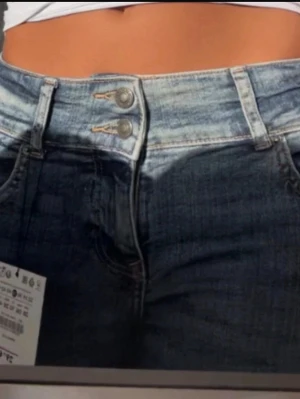 Blå bootcut jeans från Bershka - Säljer ett par blå bootcut jeans från Bershka med dubbla knappar i midjan och klassisk femficksdesign. Jeansen har slitningar framtill och är tillverkade i denim med lätt stretch. Perfekta för en avslappnad och trendig look.