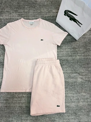 Ljusrosa loungewear-set från Lacoste - Säljer ett ljusrosa loungewear-set från Lacoste med t-shirt och shorts. Setet har den klassiska gröna krokodilloggan broderad på både tröjan och shortsen. Tillverkat i mjuk bomull för maximal komfort och avslappnad stil.
