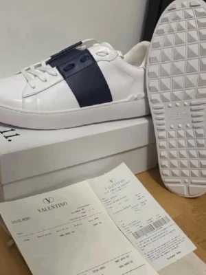 Valentino vita sneakers med blå detalj - Snygga vita sneakers från Valentino med en bred mörkblå remsa över sidan och matchande blå detaljer vid snörningen. Skorna är låga och har en klassisk siluett med vit sula och ovandel i läder. Perfekta för dig som vill ha en stilren och exklusiv look.