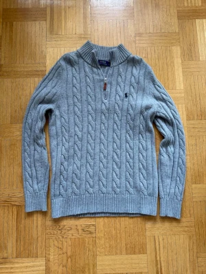 Grå kabelstickad tröja Polo Ralph Lauren - Kabelstickad grå tröja från Polo Ralph Lauren med halv dragkedja och broderad logga på bröstet. Tröjan har ribbade muddar och hög krage, perfekt för lager-på-lager. Klassisk och stilren design i mjukt material. Använt varsamt. Storlek L. Skriv om ni har några funderingar ☺️