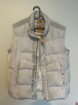 Ljusgrå dunväst från Parajumpers - Snygg ljusgrå dunväst från Parajumpers Masterpiece Series. Västen har hög krage, dragkedja och tryckknappar framtill samt två stora fickor med knappstängning. Tillverkad i ett glansigt material med quiltad design och ribbade axelpartier. Perfekt för lager på lager.