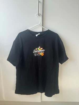 Svart Sakamoto Days t-shirt med tryck - Cool svart t-shirt med färgglatt Sakamoto Days-tryck både fram och bak. T-shirten har rund hals och korta ärmar. Perfekt för dig som gillar manga och streetwear. Materialet är mjuk bomull som känns skönt mot huden.