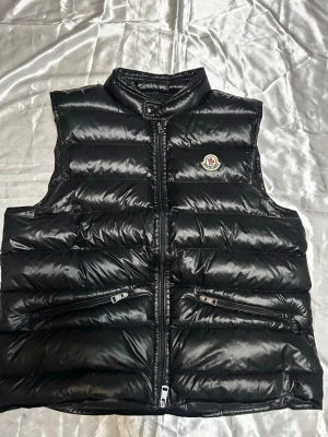 Svart dunväst från Moncler - Snygg svart dunväst från Moncler med glansig finish och klassisk logga på bröstet. Västen har hög krage, två dragkedjeförsedda fickor och quiltad design. Perfekt för lager-på-lager och streetstyle.