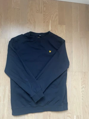  sweatshirt från Lyle & Scott - Klassisk svart sweatshirt från Lyle & Scott med rund halsringning och den ikoniska gula fågel-loggan broderad på bröstet. Tröjan har långa ärmar och ribbade muddar vid ärmslut och nederkant. Perfekt för en avslappnad och stilren look.