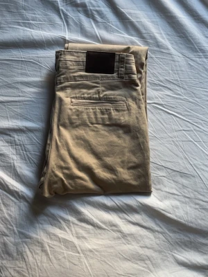 Beige chinos med bakficka - Säljer ett par klassiska beige chinos med en snygg bakficka och mörkbrunt märke på linningen. Byxorna har rak passform och är tillverkade i ett mjukt bomullsmaterial som är skönt att bära. Perfekt för dig som gillar stilrena och enkla plagg. Använda en gång 