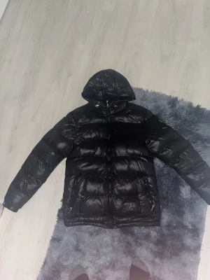 Svart pufferjacka från Alessandro - Snygg svart pufferjacka från Alessandro med huva och dragkedja framtill. Jackan har stora fickor och en diskret logga på bröstet. Tillverkad i glansigt material som ger en modern look och håller dig varm under kalla dagar.