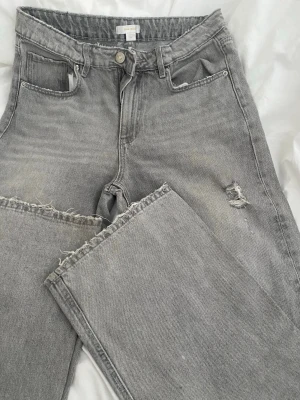 Gråa raka jeans från Gina Tricot - Säljer ett par gråa jeans från Gina Tricot young med raka ben och råa kanter. Jeansen har en liten slitning på ena låret och klassisk femficksdesign. Materialet är denim i bomull och de har hög midja för en trendig look.