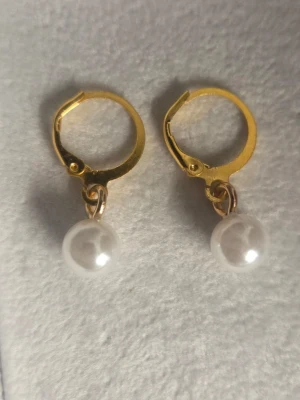 Guldfärgade örhängen med pärlor - Snygga örhängen i guldfärgat rostfritt stål med vita pärlor som hänger ned. Klassisk rund form på både ring och pärla, perfekt för dig som gillar stilrena accessoarer med en touch av elegans.