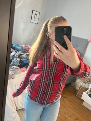 Röd rutig skjorta med knappar - Säljer en röd rutig skjorta med blå och vita detaljer. Skjortan har långa ärmar, klassisk krage och knäppning framtill. Perfekt till jeans för en avslappnad look. Materialet känns som bomull och skjortan har en normal passform. Säljer även jeansen! 