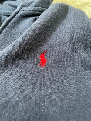 Mörkblå hoodie från Polo Ralph Lauren - Mörkblå hoodie från Polo Ralph Lauren med klassisk röd broderad logga på bröstet. Hoodien har huva och är tillverkad i mjukt bomullsmaterial som känns skönt mot huden. Perfekt för en avslappnad och stilren look.