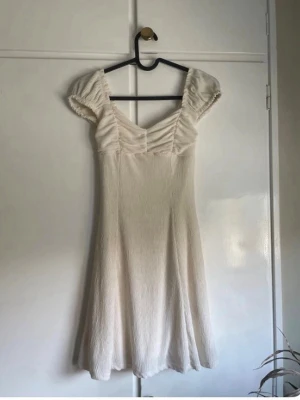 Ljus kortklänning med puffärm - Söt ljus kortklänning med puffärmar och v-ringad hals. Klänningen har en lätt rynkad överdel och är tillverkad i ett mjukt, strukturerat tyg. Perfekt för varma dagar och ger en romantisk vibe.