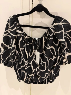 Ny Blus Svartvit blus med abstrakt mönster - Trendig svartvit blus från MQ  med abstrakt mönster. Modellen har puffiga korta ärmar, rundad halsringning och croppad passform. Materialet är lätt och har en plisserad struktur som ger extra volym och stil.Helt ny med lappar på 