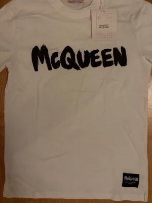 Vit McQueen t-shirt med tryck - Snygg vit t-shirt från Alexander McQueen med stort svart McQueen-tryck på bröstet och en liten patch nertill. Klassisk rund hals och korta ärmar. Perfekt för dig som gillar streetstyle och vill ha något från ett ikoniskt märke.