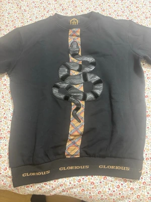 Svart t-shirt med orm från Glorious - Svart t-shirt från Glorious med korta ärmar och ett stort ormtryck framtill. Snyggt rutigt band i mitten och loggan 'GLORIOUS' i guld på mudden. Perfekt för dig som vill sticka ut med coola detaljer och unik design.