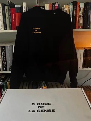 Mörkblå sweatshirt D'ONCE DE LA SENSE - Svart sweatshirt med gul text 'D'ONCE DE LA SENSE' på bröstet. Klassisk rund halsringning och ribbade muddar vid ärmslut och nederkant. Perfekt för en avslappnad och stilren look.