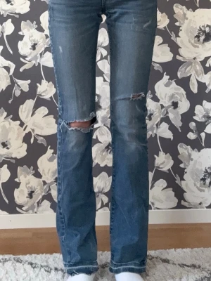 Blå bootcut jeans med slitningar - Säljer ett par blå bootcut jeans med slitningar på knäna och lätt tvättad look. Jeansen har klassisk femficksdesign och råa kanter vid bensluten. Perfekta för en avslappnad och trendig stil. Jag är 173 och de passar perfekt! 