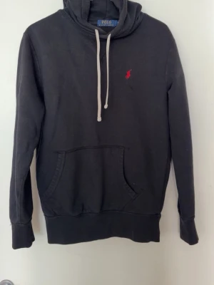 Svart hoodie från Polo Ralph Lauren - Svart hoodie från Polo Ralph Lauren med klassisk röd logga broderad på bröstet. Hoodien har en stor magficka framtill. Tillverkad i mjukt bomullsmaterial och har ribbade muddar vid ärmslut och nederkant.