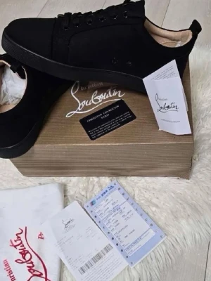 Loubs  - Stilrena svarta sneakers från Christian Louboutin med klassisk röd sula. Skorna har lågt skaft, svarta skosnören och ovandel i mocka. Insidan är beige och designen är minimalistisk men lyxig, perfekt för dig som gillar exklusiva detaljer.