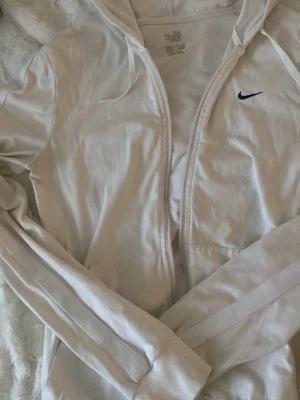 Vit zip-hoodie från Nike - Snygg vit hoodie från Nike med dragkedja framtill och klassisk huva. Diskret Nike-logga i svart på bröstet. Tillverkad i mjukt material och har långa ärmar samt ribbade muddar. Perfekt för en sportig och avslappnad stil.