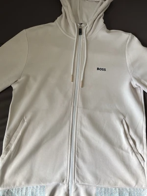 Vit zip hoodie från BOSS - Snygg vit hoodie från BOSS med dragkedja framtill och justerbar huva med snören. Diskret BOSS-logga på bröstet och två fickor på sidorna. Tillverkad i mjukt material som känns skönt mot huden. Perfekt för en clean och stilren look.