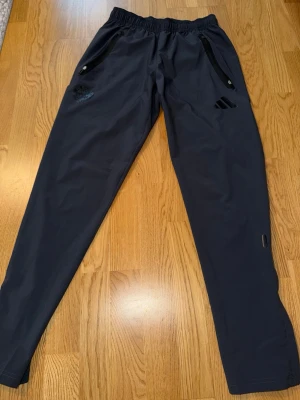 Svarta träningsbyxor från Adidas - Säljer ett par svarta träningsbyxor från Adidas med regular fit. Byxorna har resår i midjan med dragsko, två fickor med dragkedja och diskret Adidas-logga på låret. Perfekta för gymmet eller löprundan.