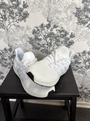 Adidas Ultraboost vita sneakers - Vita Adidas Ultraboost sneakers med stickad mesh-ovandel, klassiska tre ränder på sidan och tjock, responsiv Boost-sula. Skorna har vita snören och svarta detaljer på sulans fram- och bakkant. Perfekt för dig som gillar sportig och clean stil.