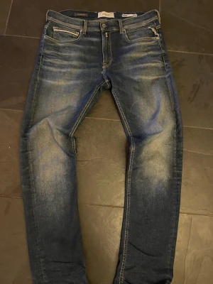 Blå slim fit jeans från Replay - Säljer ett par blå slim fit jeans från Replay med snygga slitningar och tvättade detaljer. Klassisk femficksmodell med dragkedja och knapp i midjan. Jeansen har en modern passform och är perfekta för en avslappnad stil. Mått: Midja 44cm, benlängd: 104cm, innerbenlängd: 79cm, Benöppning: 18cm. 