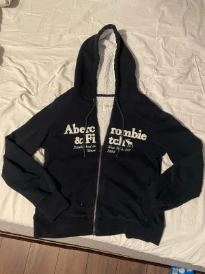 Svart hoodie Abercrombie & Fitch - Mysig svart hoodie från Abercrombie & Fitch Soft A&F Collection med vit teddyfoder i huvan och dragkedja framtill. Stor logga i vitt på bröstet och fickor på sidorna. 