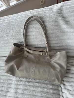 Beige handväska med dubbla handtag - Stilren beige handväska i mjukt konstläder med dubbla långa handtag och enkel guldfärgad detalj. Väskan har en avlång, rymlig form och minimalistisk design som passar till många olika looks.