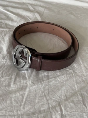 Brunt Gucci skärp med silverfärgat spänne - Snyggt brunt skärp från Gucci med ett stort, rundat silverfärgat GG-spänne. Bältet är gjort i slätt skinn och har flera hål för justering. Perfekt accessoar för att lyfta vilken outfit som helst.