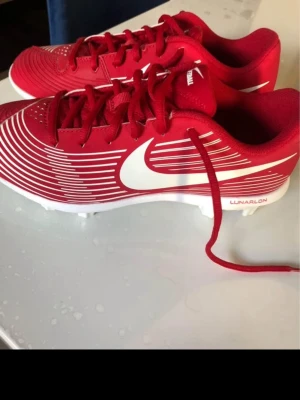 Röda Nike fotbollsskor Lunarlon - Säljer ett par röda Nike fotbollsskor med vita detaljer och stora vita swoosh-loggor på sidorna. Skorna har snörning, plastdobbar och texten 'Lunarlon' på sidan. Tillverkade i syntetmaterial med modern design och mönstrade linjer för extra stil.