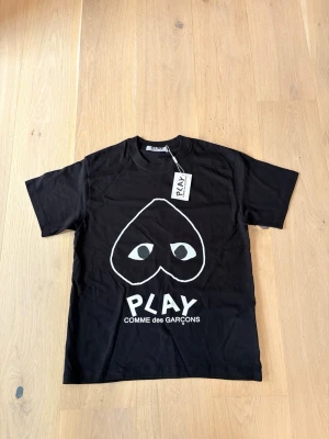 Svart Comme des Garçons PLAY t-shirt - Svart t-shirt från Comme des Garçons PLAY med det ikoniska hjärtat med ögon tryckt framtill. Klassisk passform och rund hals.  Perfekt för dig som gillar streetwear och stilrena prints.                      Tror du vet varför det billiga priset😊