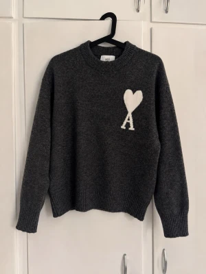 Mörkgrå stickad tröja från Ami - Mysig mörkgrå stickad tröja från Ami med ett vitt hjärta och bokstaven 'A' broderat på bröstet. Tröjan har rund halsringning, långa ärmar och ribbade muddar vid ärmslut och nederkant. Perfekt för kyliga dagar och enkel att styla.