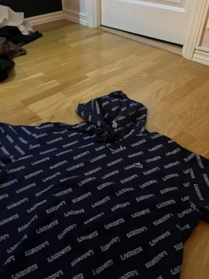 Marinblå Lacoste LIVE hoodie M - Säljer en marinblå hoodie från Lacoste LIVE i storlek M. Hoodien har ett all-over print med Lacoste-loggan och den klassiska krokodilen broderad på bröstet. Utrustad med huva och dragsko. Perfekt för dig som gillar streetwear och vill sticka ut.