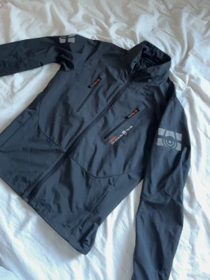 Svart vindjacka från Sail Racing - Svart vindjacka från Sail Racing med dragkedja framtill, två sidofickor och en bröstficka med röda detaljer. Jackan har tryckta loggor på ärmen och bröstet samt hög krage. Tillverkad i lätt och slitstarkt material, perfekt för blåsiga dagar.