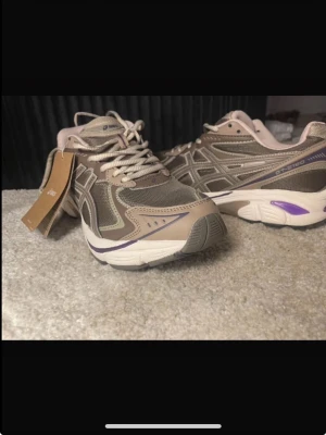 Beige, brun och lila Asics Gel Kahana sneakers - Snygga sneakers från Asics i beige och bruna toner med lila detaljer. Skorna har en chunky sula i vitt och meshpaneler för extra andningsförmåga. Klassisk snörning och retroinspirerad design som passar perfekt till streetwear-looken. Skorna är oanvända och helt i nyskick, prislappen är avtagen men sulorna är helt rena och inga tecken på användning!