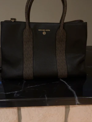 Svart handväska från Michael Kors - Snygg svart handväska från Michael Kors med bruna detaljer och logotyp på framsidan. Väskan har både handtag och en justerbar axelrem i brunt med MK-mönster. Tillverkad i strukturerat konstläder och har guldfärgade metalldetaljer.