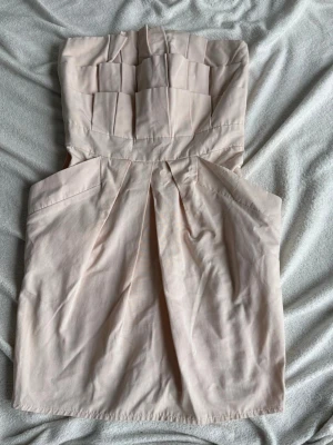 Ljusbeige axelbandslös kort klänning - Säljer en stilren axelbandslös klänning i ljusbeige med veckad överdel och markerad midja. Klänningen har en kort längd. Perfekt för dig som gillar minimalistisk och elegant stil.