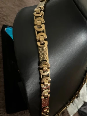 Guldigt kedjehalsband i metall - Snyggt halsband i guldfärgad metall med unika, rektangulära länkar och detaljerade mönster på vissa delar. Kedjan har en robust och modern design som sticker ut och passar perfekt till dig som gillar statement-smycken.