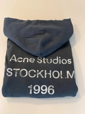 Mörkblå hoodie Acne Studios - Snygg mörkblå hoodie från Acne Studios med tryck i vitt på ryggen där det står 'Acne Studios STOCKHOLM 1996'. Klassisk passform med huva och ribbade muddar. Tillverkad i mjuk bomull för maximal komfort.