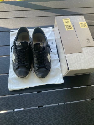 Svarta Golden Goose Superstar sneakers - Golden Goose Superstar sneakers i svart läder med vit stjärndetalj på sidan och vita sulor. Klassisk låg modell med svarta skosnören och GGDB-märke på plösen. Snygg streetstil som passar till det mesta och ger en cool vibe.