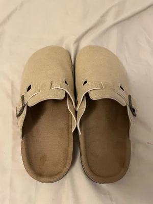 Beige mockatofflor med spänne - Säljer ett par snygga beige tofflor i mocka med justerbart metallspänne på sidan. De har en klassisk slip-in design och svart sula som ger bra grepp. Perfekta för dig som gillar stilrena och bekväma skor.