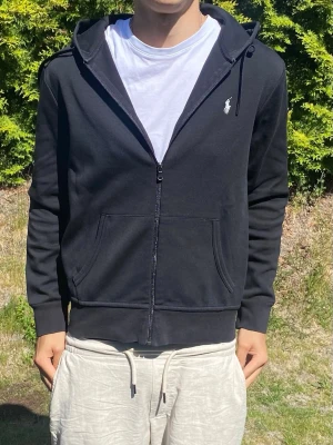 Svart hoodie från Polo Ralph Lauren - Snygg svart hoodie från Polo Ralph Lauren med dragkedja, huva med dragsko och två stora fickor framtill. Klassisk vit logga på bröstet. Tillverkad i mjukt bomullsmaterial som känns skönt mot huden. Perfekt för en avslappnad och stilren look.