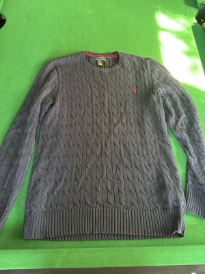 Mörkgrå kabelstickad tröja Polo Ralph Lauren - Kabelstickad marinblå tröja från Polo Ralph Lauren. Säljer då den inte används. Den är i storlek barn XL men sitter som en S. Hör av dig vid fler frågor